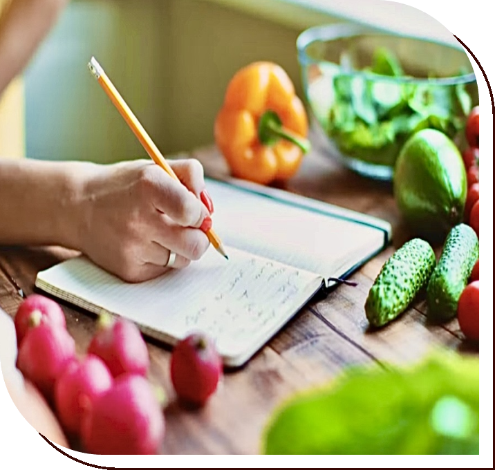 A person writing on a notebook with vegetables around it.